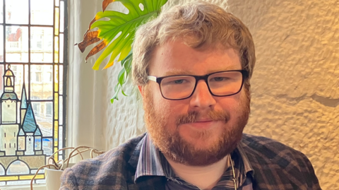 A man with ginger hair, glasses and a beard in a blue shirt in front of a small stained glass window