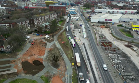 An aerial view of a construction site beside a major road. On the left, apartment buildings and trees surround areas marked with orange fencing. On the right, vehicles travel along the road alongside construction equipment, cones and barriers. Commercial buildings are in the background.