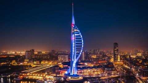 Aerial night view of the illuminated Spinnaker Tower in Portsmouth, glowing in blue and red lights with reflections shimmering on the water and surrounding city buildings lit up in the background.
