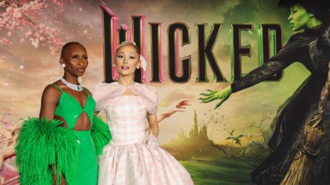 Cynthia Erivo wears a bright green dress. She is standing next to Ariana Grande who is wearing a light pick ginham dress. They stand in from of the Wicked poster image