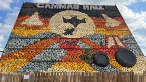 More than 3,500 pumpkins have been put together in a mosaic, which is standing vertical, at a farm. The pumpkins are all different colours. The mosaic is large and shows a tractor and fields. 