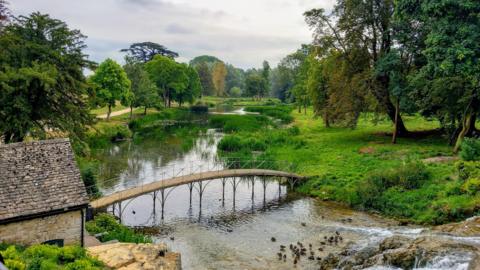 A river runs through the centre of the picture, there is a humped footbridge over the river and on either side the banks are full of lush green vegetation and trees. There is a small grey stone building at one end of the bridge. And there is a small flock of brown birds in the water.