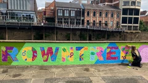 A view of a vibrant mural, painted along a low brick wall along a riverside path. The mural depicts the word 'Rowntree' painted in bright multicolours against predominately lime-green background. Each letter also contains leaf and flower motifs within it. On the opposite side of the river, a metal walkway and several buildings housing bars can be seen. In the corner, a woman with long blonde hair and wearing black paints onto the mural.