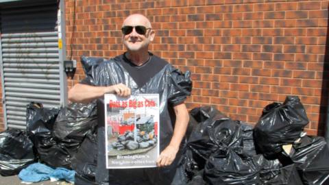 Kevin is bald and wearing black sunglasses, he is wearing a black t-shirt with a bin bag fashioned into a top, over the top of it. He is holding a calendar that says "rats as big as cats". In the background behind him there is a pile of black bin bag with rubbish scattered on the pavement