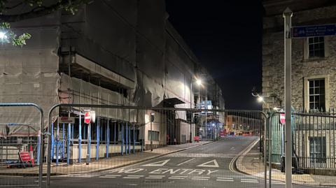A picture of fences in front of the building. There is a large building behind it covered in scaffolding and sheets.