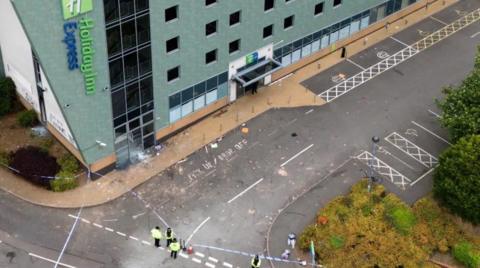 An aerial view of the Holiday Inn Express hotel with a police cordon. There are officers stood by police tape, with damage to a section of the building visible.