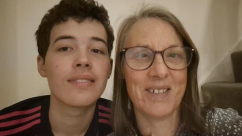 A woman with brown hair and glasses sat on a staircase with a white wall. Also in the close-up selfie is her teenage son, who has black hair and is wearing a black sports top. Both people are smiling.