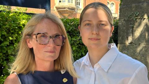 A woman wearing a dark blue dress is standing next to a girl wearing a white shirt. They are next to a hedge and a stone wall. Further behind them is a red brick building with stone window surrounds and elaborate bay windows.