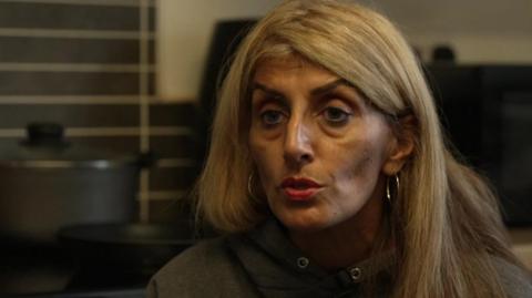 Farah McLaren, who has long blonde hair and is wearing a grey Nike branded sweatshirt, speaks to a tv reporter with her kitchen in the background