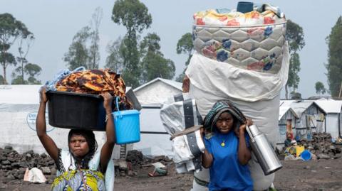 Two women flee a camp for displaced people with their belongings. They carry a mattress, a large thermos flash and more.