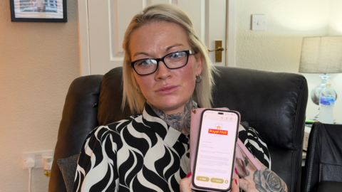 Samantha Fisher wearing black-rimmed glasses and a black and white patterned shirt, is sitting on a black recliner sofa and is holding up her pink and black phone. On the screen is an email from Royal Mail, showing the logo at the top, followed by text which apologises for the ongoing delays she is facing. In the background is a white door, a lamp and a double plug socket.
