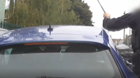 The back of a blue car pictured from immediately behind. A police officer is stood alongside it with his arm raised, he is holding a baton in his left hand which he is about to smash into the window. 