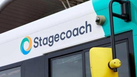 The right side of a white and mint green bus with a yellow wing mirror. The image is zoomed in above the driver's window showing the top of the window and the word Stagecoach written in black letters on the bus itself