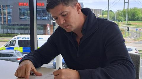 Luke Campbell with short, dark hair wearing a blue top signing the acceptance of office declaration in an glass sided office with a view of the Humber Bridge in the background