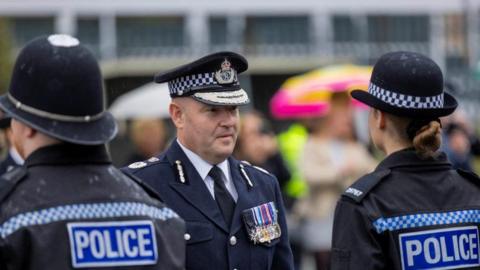 West Midlands Police Chief Constable Craig Guildford at a passing out parade, wearing full uniform and medals on his chest. He is facing two young officers 