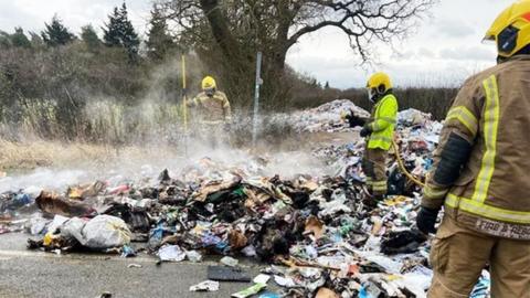 Firefighters use a hose to tackle a smoking pile of rubbish dumped on the road from a bin lorry