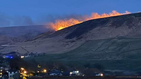 Large orange flames line the crest of a hill marked with patches of brown vegetation, in the foreground are a number of houses.