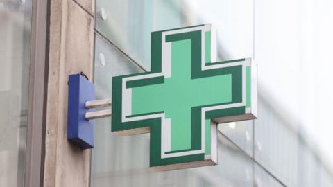 A pharmacy sign of a green plus sign on a shop wall with glass screens behind