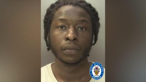 A mugshot of Elie Mey wearing a white t shirt. He has black braided hair and has a short black beard