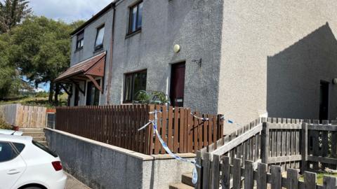 The exterior of a semi-detached property in Bonar Bridge. There is blue and white police taped across the front steps to the property.