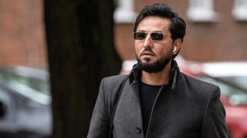 Image shows Salwan Momika wearing sunglasses, a grey coat and airpods