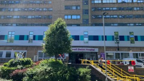 Outside of Yeovil district hospital's main entrance, with yellow steps and green bushes. The building is brown bricks