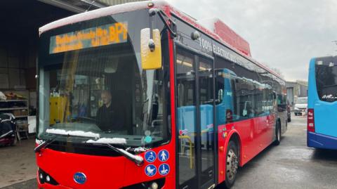 A red single-decker electric bus stands in a depot.