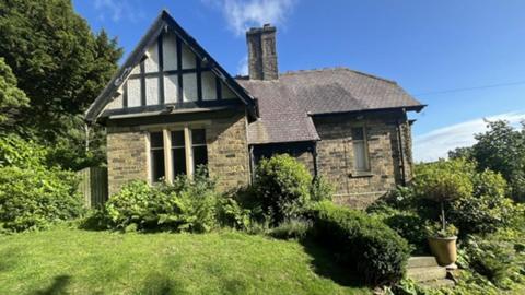 A detached stone building with a tall roof apex and chimneys, surrounded by lawn and shrubberies.