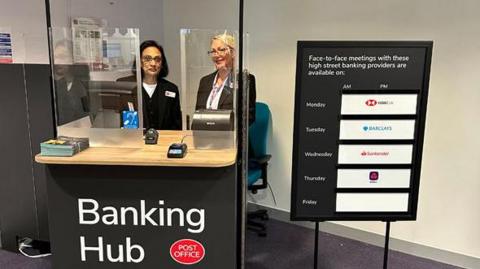 Two people sat behind clear screens at a stand with a sign reading "Banking Hub".