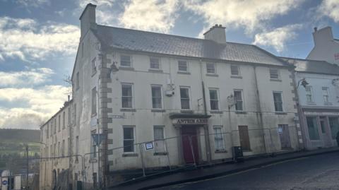 A white building with a red door and red lettering above the door which reads "Antrim Arms". The building is three stories high and is showing signs of wear. Two windows are boarded up and there is a plant growing out of the guttering. The building is surrounded by a metal railings