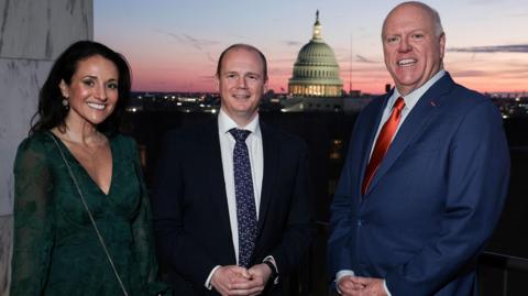 Three people in formal attire pose for a picture at sunset with Capitol Hill visible in the background. The woman on the left has black hair and is wearing a purse which says 'America 250'. The man in the middle is wearing a dark suit, the man to his right a blue suit.
