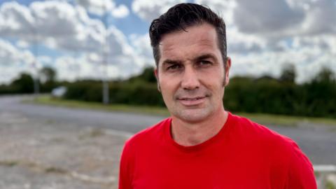 Billy Crotty, wearing a red T-shirt, looks directly into the camera. He has dark, slicked-back hair. Behind him, slightly blurred, is an empty road.