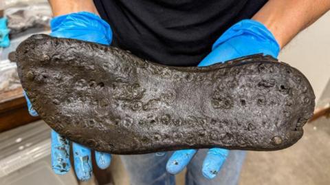 The sole of a large black shoe being held by a person wearing blue gloves.