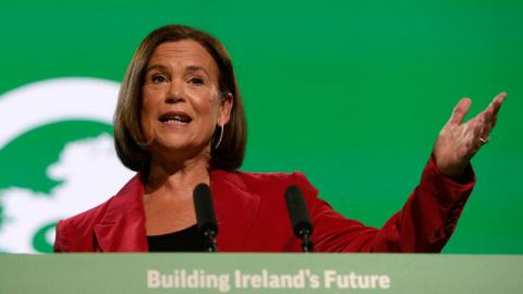 Mary Lou McDonald is pictured speaking at her party's annual conference. She has shoulder length brown hair and is dressed in a red blazer and back top, gesturing with her left hand. Behind her the backdrop is bright green and on the podium in front of her are the words 'Building Ireland's future'.
