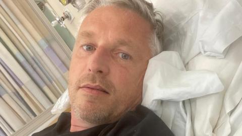 A close up of Richard Walker, lying in a hospital bed in a black T-shirt