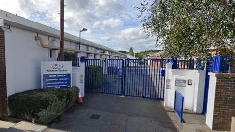 The gated entrance to Martin-Baker site on Lower Road. There are tall blue gates across the entrance and a sign to their left with instructions for visitors. A long white one-storey building to the left with further buildings beyond the gates, and a tree on the right. 