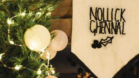 White baubles on a green Christmas tree with an embroidered white banner that says Nollick Ghennal above a holly leaf and berries.