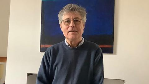 Peter is standing in a living room with a painting behind him of a night sky. He is wearing a blue pullover and glasses