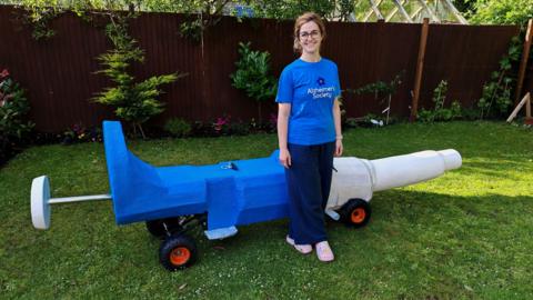 Inês Rodrigues in a blue Alzheimer's Society t-shirt stood in front of the pipet shaped soap box which is blue and white.