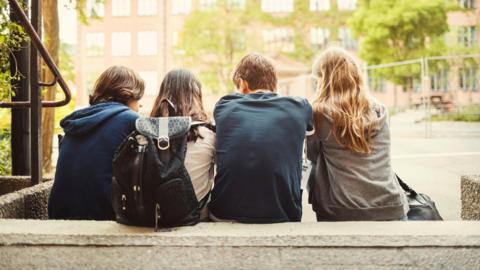Rear view of four teenagers sitting on steps outdoors. There are buildings in the background.