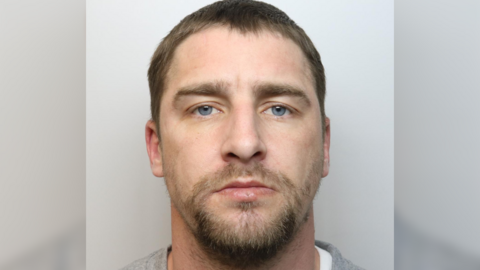 A police custody image of Matthew Brown. He has short brown hair and a short beard and is wearing a light-grey jumper and white t-shirt.