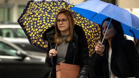 Danielle Sassoon, carrying a brown file folder and holding an umbrella with flowers. She has a neutral expression on her face and is in casual clothing. An unidentified woman walks beside her. 