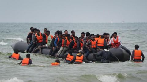 A large group sitting on an inflatable small boat, many sitting astride the outer edges. Six people are seen wading in deep sea water towards the boat. Most, but not all, are wearing orange life-jackets.