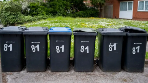 A row of six black, numbered wheelie bins on a pavement in front of a grass lawn and a house