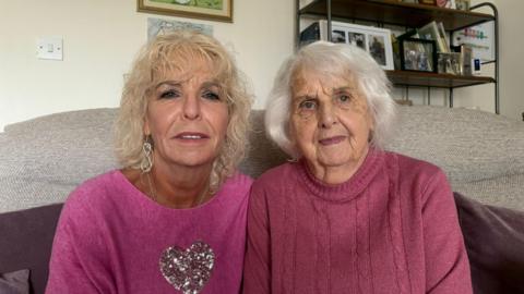 Judith Bosustow sitting on the left with blond hair in a pink jumper, beside her mum, Joan, in a purple jumper with white hair