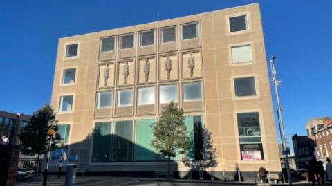 The exterior of Grimsby Central Library. It is a five-storey brutalist concrete building with double-sized windows to the ground and first floor, It is decorated with sculptures of people on one of the floors.