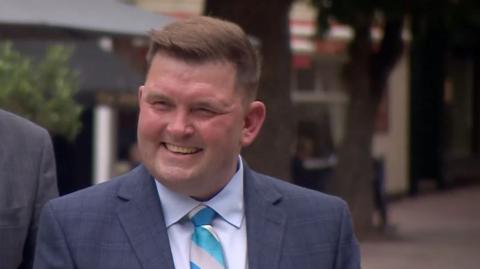 Gary Burgess who is wearing a blue suit and tie and smiling at a camera