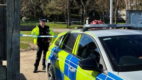 A marked fluorescent yellow and blue police car is parked outside of one of the entrances to Sparrow's Nest Gardens. Behind the car, which is parked, there is a police officer in uniform standing in an area which has been cordoned off with blue and white police tape.