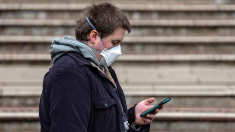 A young person wearing a hoodie, jacket and a mask across his face reads his phone.