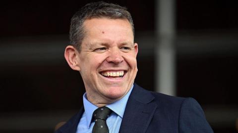 Rhun ap Iorwerth in a dark blue suit and a light blue shirt, wearing a dark green and pink dotted tie. It is a head and shoulders shot.
